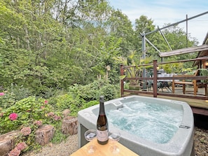 Ein Whirlpool mit einer Weinflasche und zwei Gläsern auf einem Holztisch, umgeben von üppigem Grün und einer Holzterrasse.