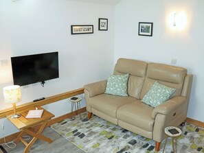Ein Wohnzimmer mit einem beigen Sofa, zwei gemusterten Kissen, einem Flachbildfernseher, einem hölzernen Beistelltisch, einer Lampe und einem gemusterten Teppich.