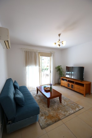 A living room with a sofa, a coffee table, a TV on a wooden stand, and a ceiling fan. - Elysia park - Charming 2BR Townhome with Pool, Gym, Free parking-Paphos central (Pafos)
