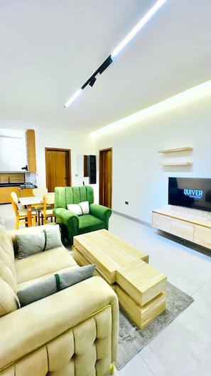 Uma sala moderna, com sofá, mesa de centro, televisão e cozinha ao fundo.