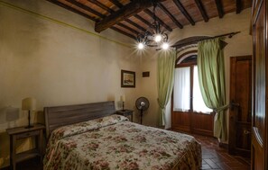 Ein Schlafzimmer mit Holzbalkendecke, einem Bett mit floralem Bettzeug, einem Nachttisch mit Lampen, einem Ventilator und einem Bild an der Wand.