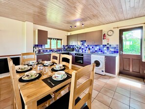 Área de refeições com mesa de madeira posta para quatro pessoas, cozinha com eletrodomésticos modernos e vista para o verde lá fora.