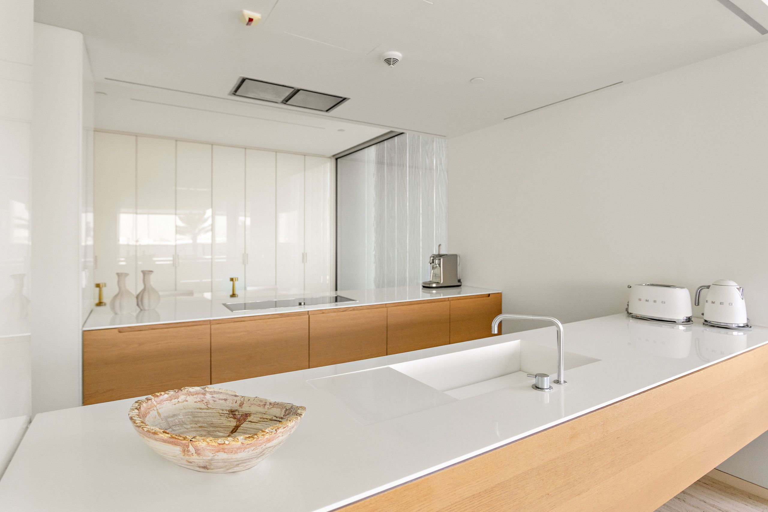 A modern kitchen with a white countertop, wooden cabinets, a large mirror, and a coffee maker.