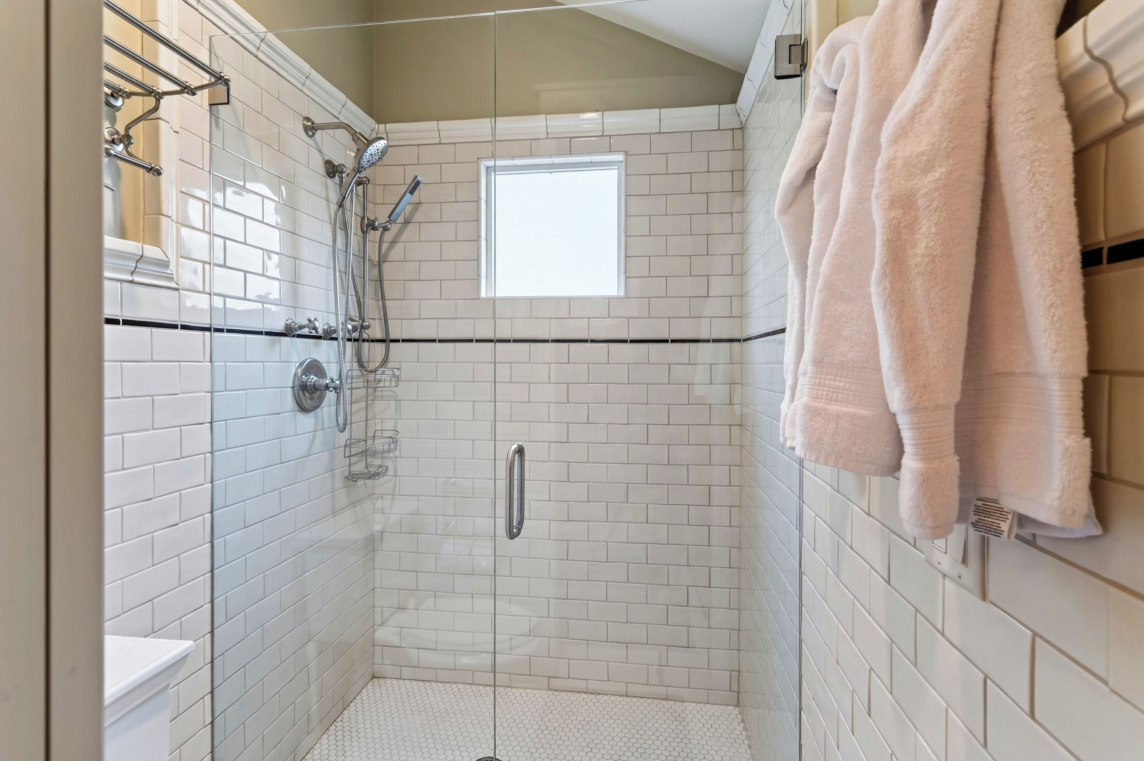 Eine Dusche ohne trittfeste Barriere mit weißen, rechteckigen Fliesen, einer Glastür und einem Handtuchhalter mit Handtüchern.