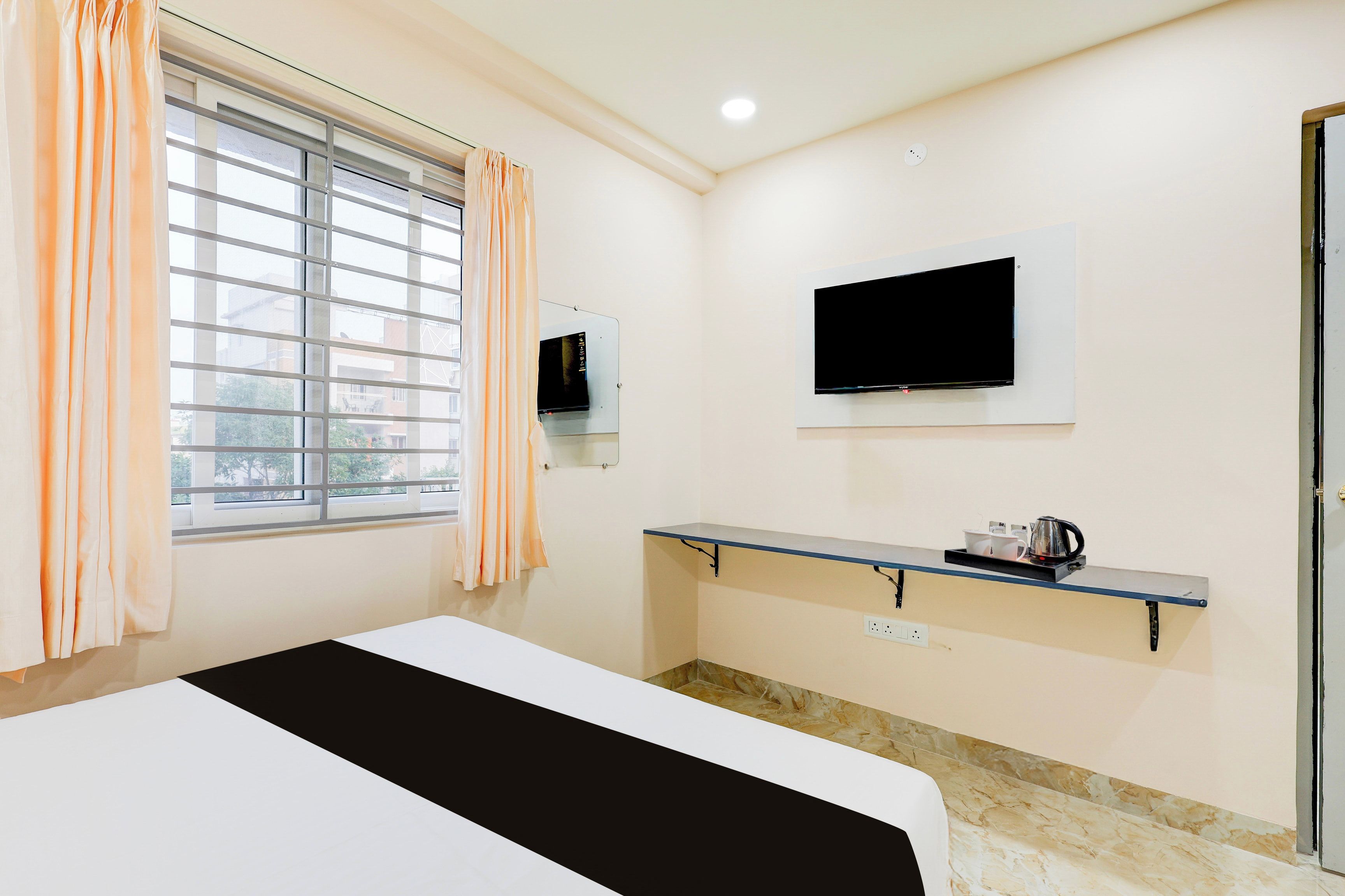 Habitación de hotel con una cama, un televisor montado en la pared, un estante con un hervidor y una ventana con cortinas.