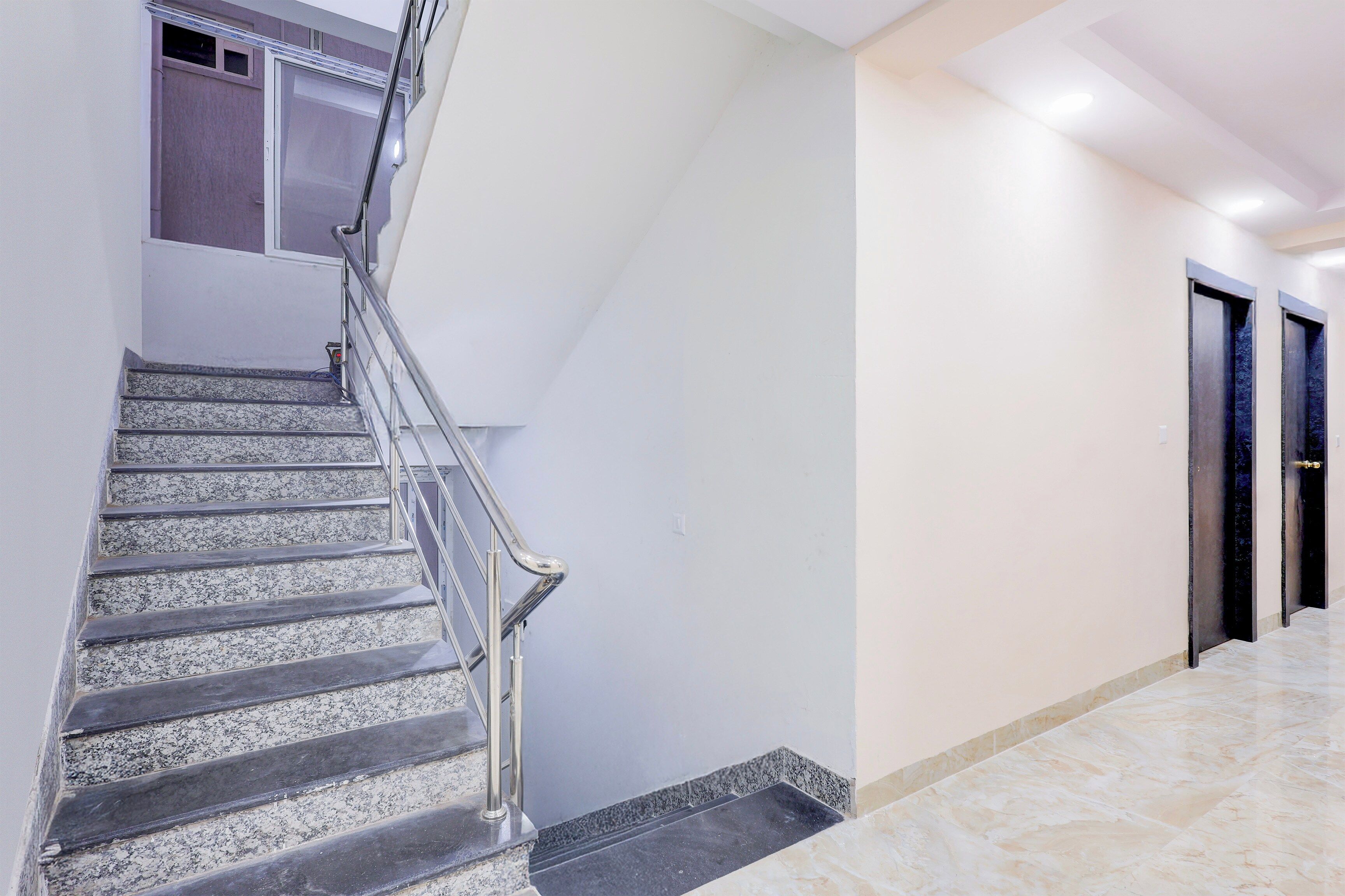 Una escalera con barandilla de metal que lleva a una plataforma, una puerta con marco oscuro y un pasillo con piso de mármol.