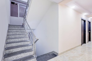 Un escalier avec une rampe métallique menant à un palier, une porte dotée d’un cadre sombre et un couloir au sol en marbre.