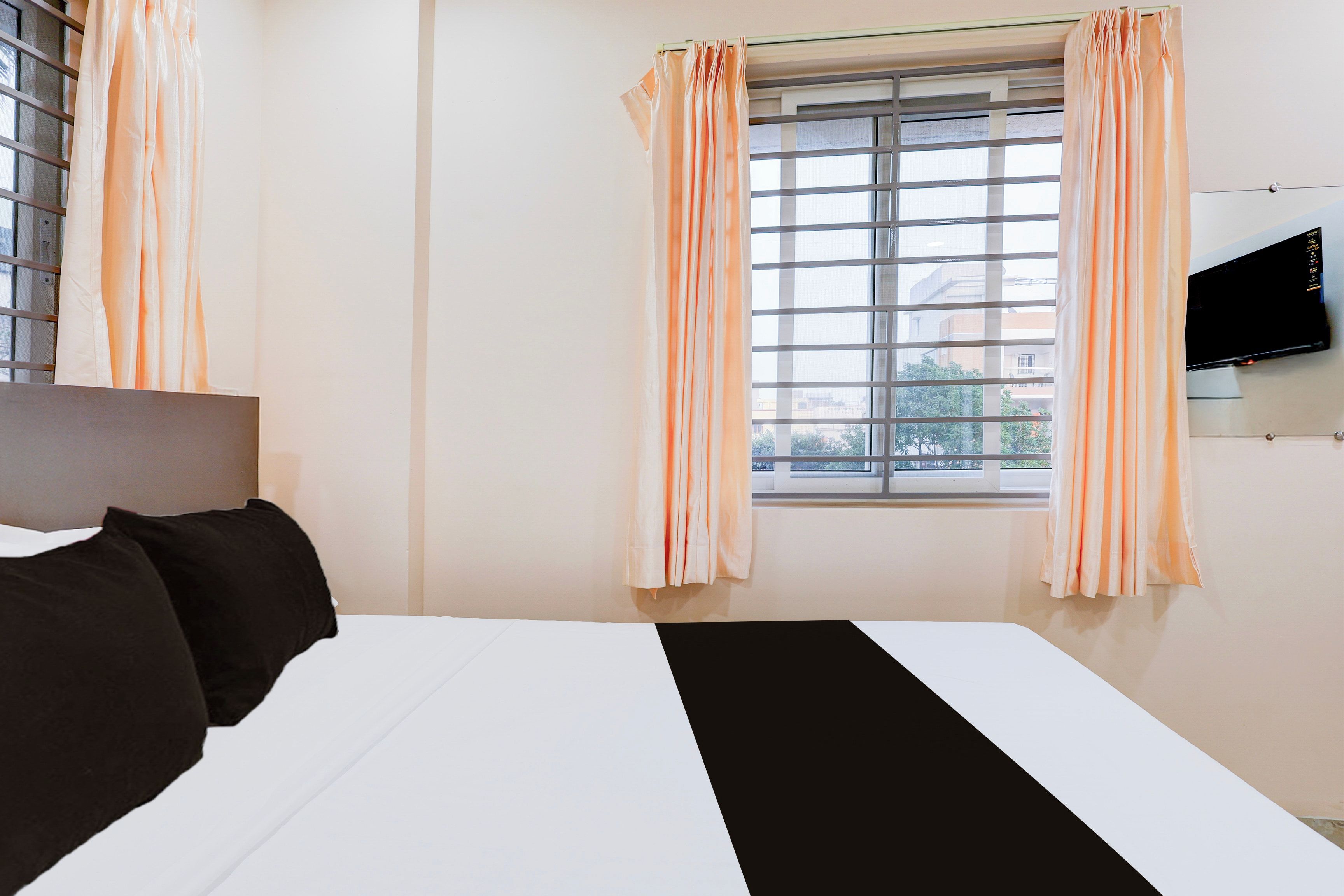 Habitación de hotel con cama, ropa de cama blanco y negro, cortinas color melocotón, una ventana con persianas y un televisor de pantalla plana montado en la pared.