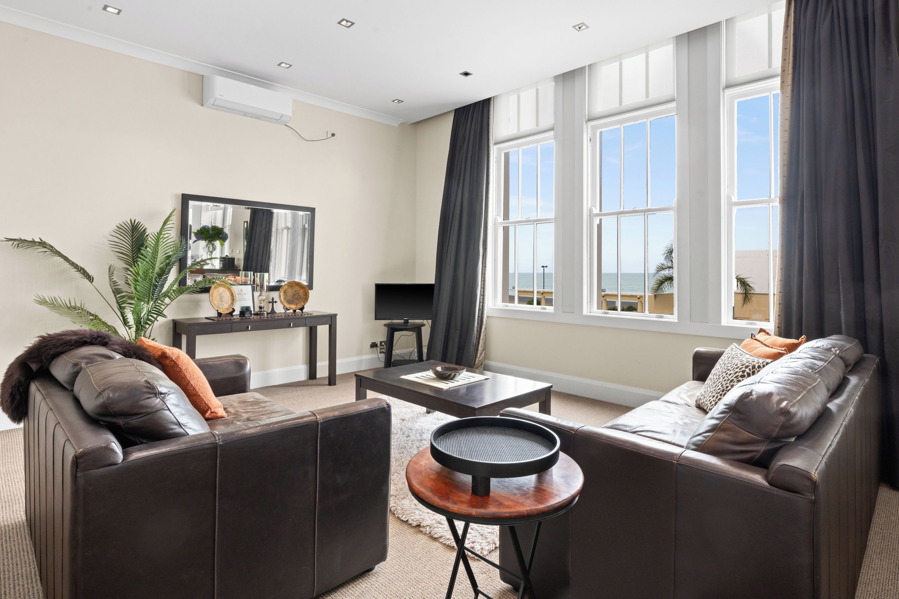 A modern living room with leather sofas, a coffee table, a TV, and a large window with a view of the sea.