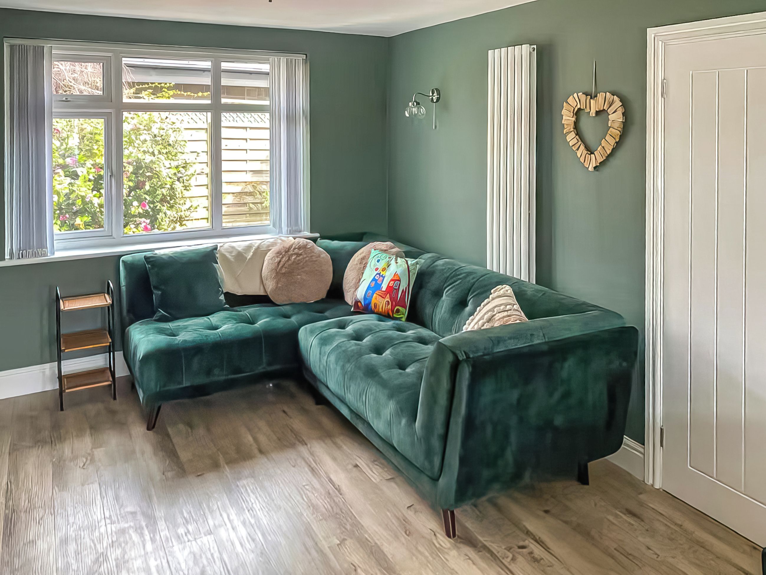Quarto com sofá de canto verde, janela com persianas, radiador e decoração em formato de coração na parede.