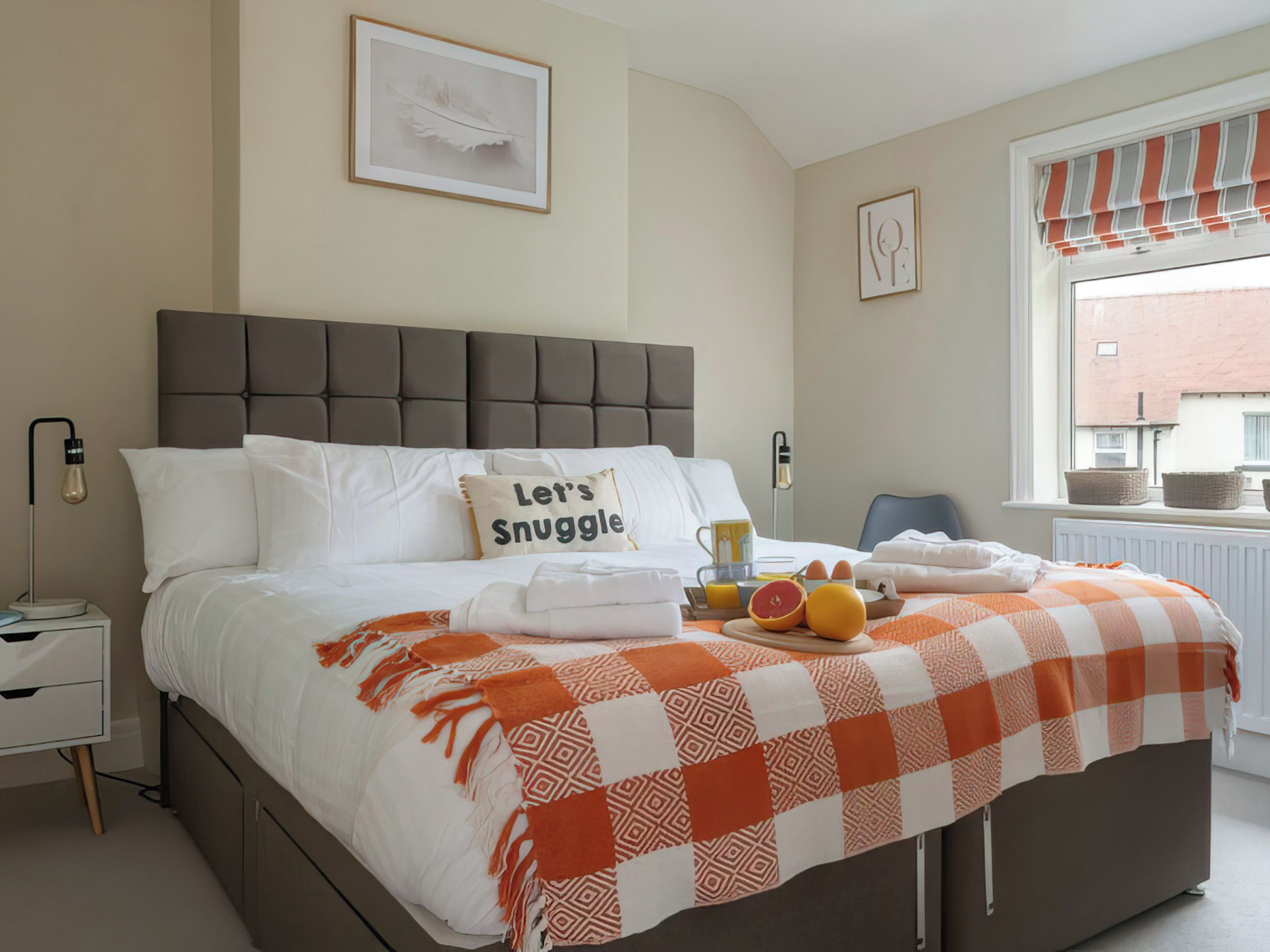 Ein ordentlich bezogenes Bett mit einer karierten Decke, ein Kopfkissen mit dem Aufdruck 'Let's Snuggle' und ein Tablett mit Frühstücksangeboten.