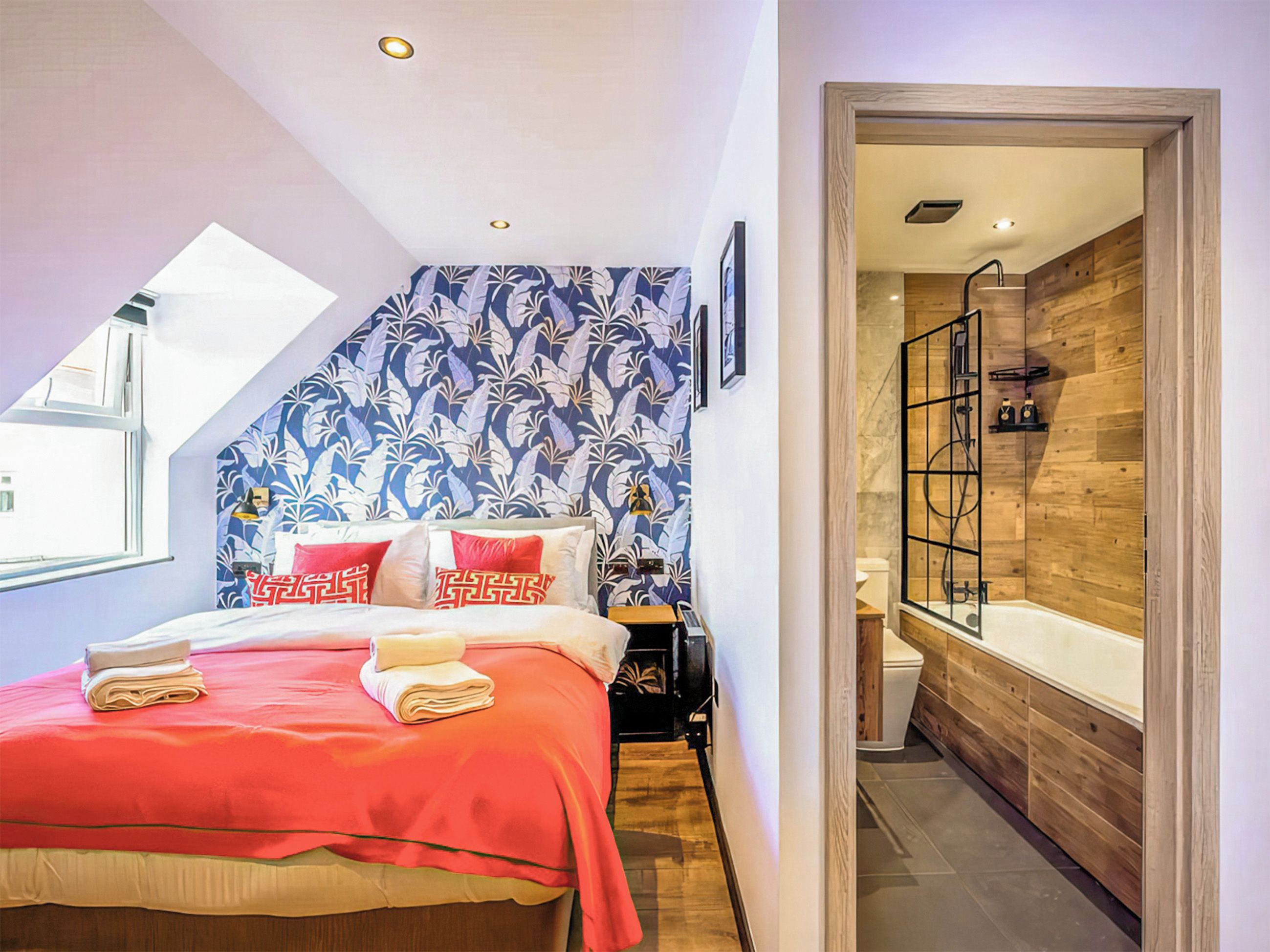 Ein Schlafzimmer mit Bett, Nachttisch, einer Wand mit Blumenmuster, einer Dusche mit Glastür und einem Oberlicht.