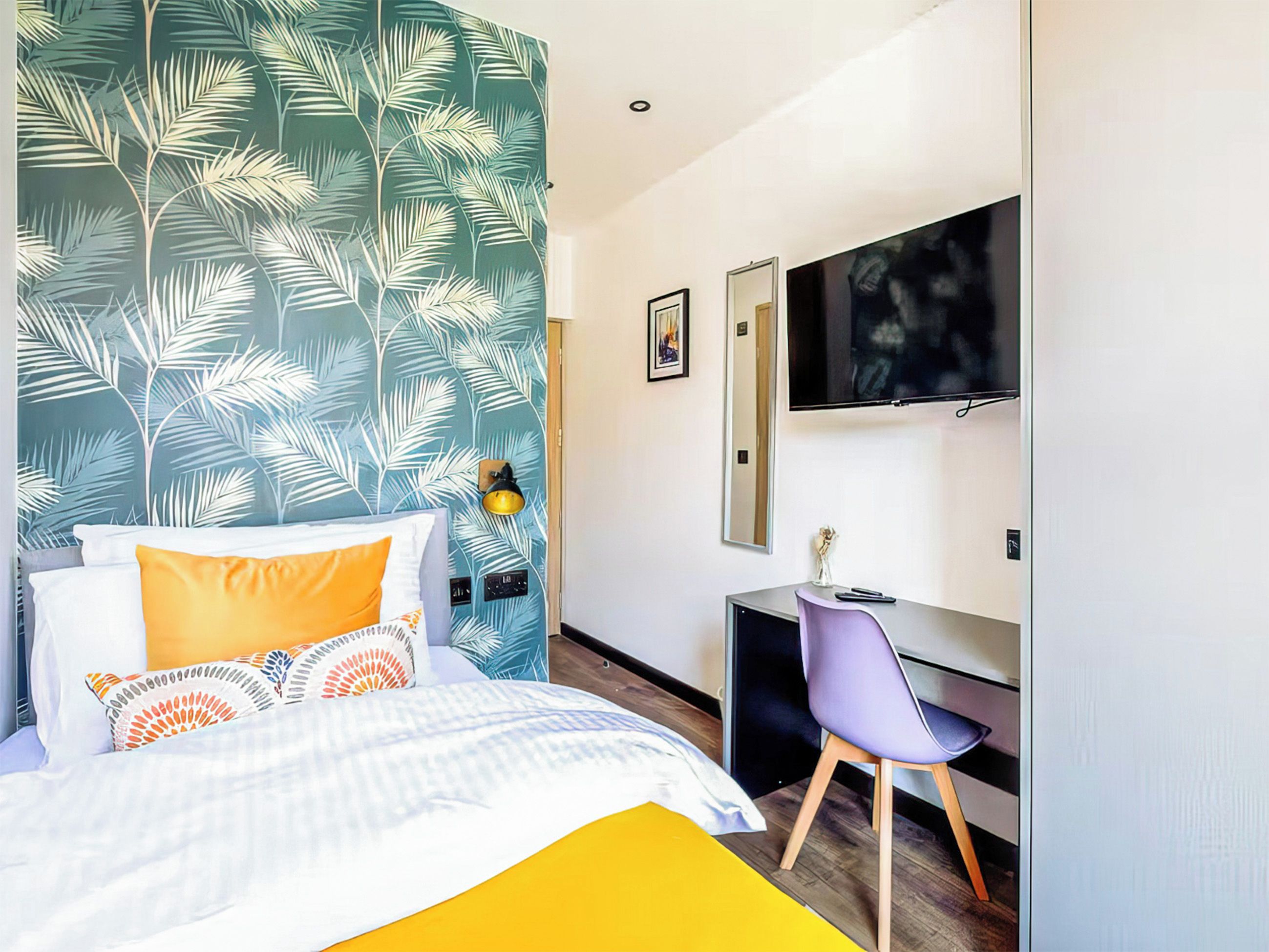 Quarto de hotel com cama, escrivaninha, cadeira, televisão e parede com desenho de folhagem.