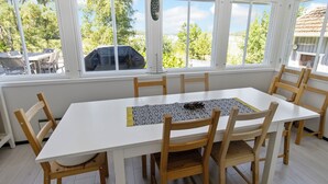 Ein Essbereich mit einem weißen Tisch und Holzstühlen, großen Fenstern mit Blick auf Bäume und ein Gebäude sowie ein dekorativer Gegenstand auf der Fensterbank.