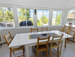 A dining area with a white table and wooden chairs, large windows with a view of trees and a building, and a decorative item on the windowsill. - Villa niemelä by Interhome (Naantali)