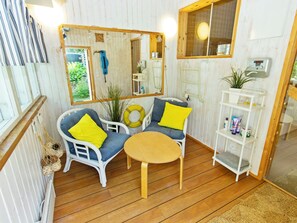 A cozy room with a wooden floor, two wicker chairs with cushions, a small round table, a mirror, a shelf with toiletries, and a window with a view of greenery.