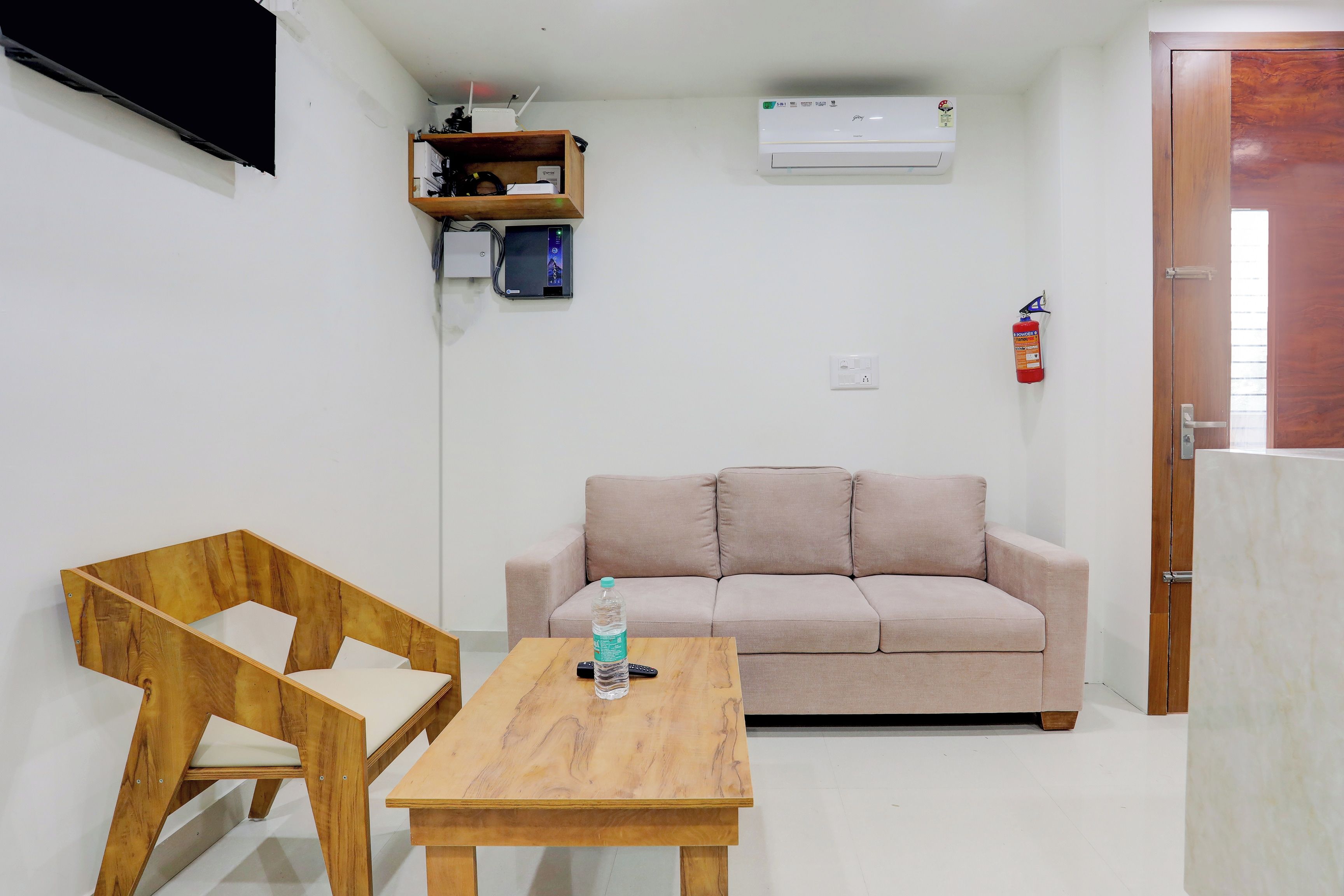 A small, well-lit room with a wooden table, a beige sofa, a television mounted on the wall, and a fire extinguisher.