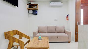 A small, well-lit room with a wooden table, a beige sofa, a television mounted on the wall, and a fire extinguisher.