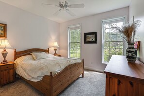 Un dormitorio con una cama de madera, dos lámparas de noche, un ventilador de techo y dos ventanas con vistas a la vegetación.
