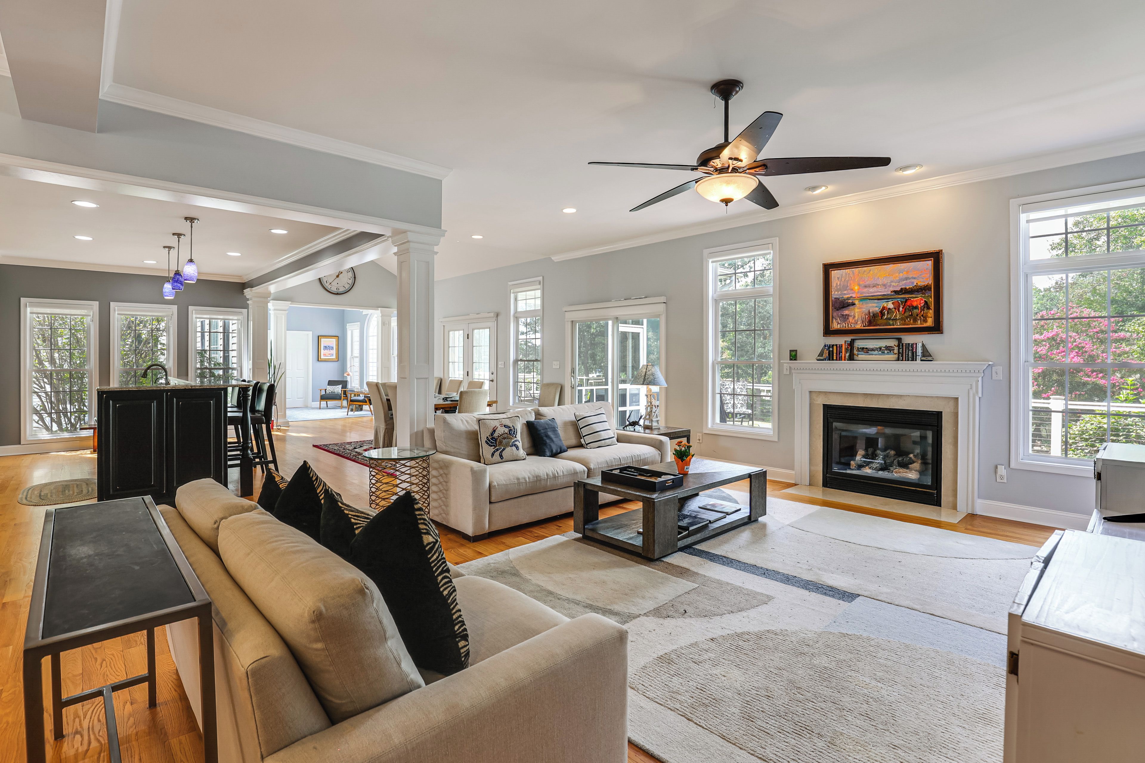 A spacious living room with a fireplace, large windows, and a ceiling fan.