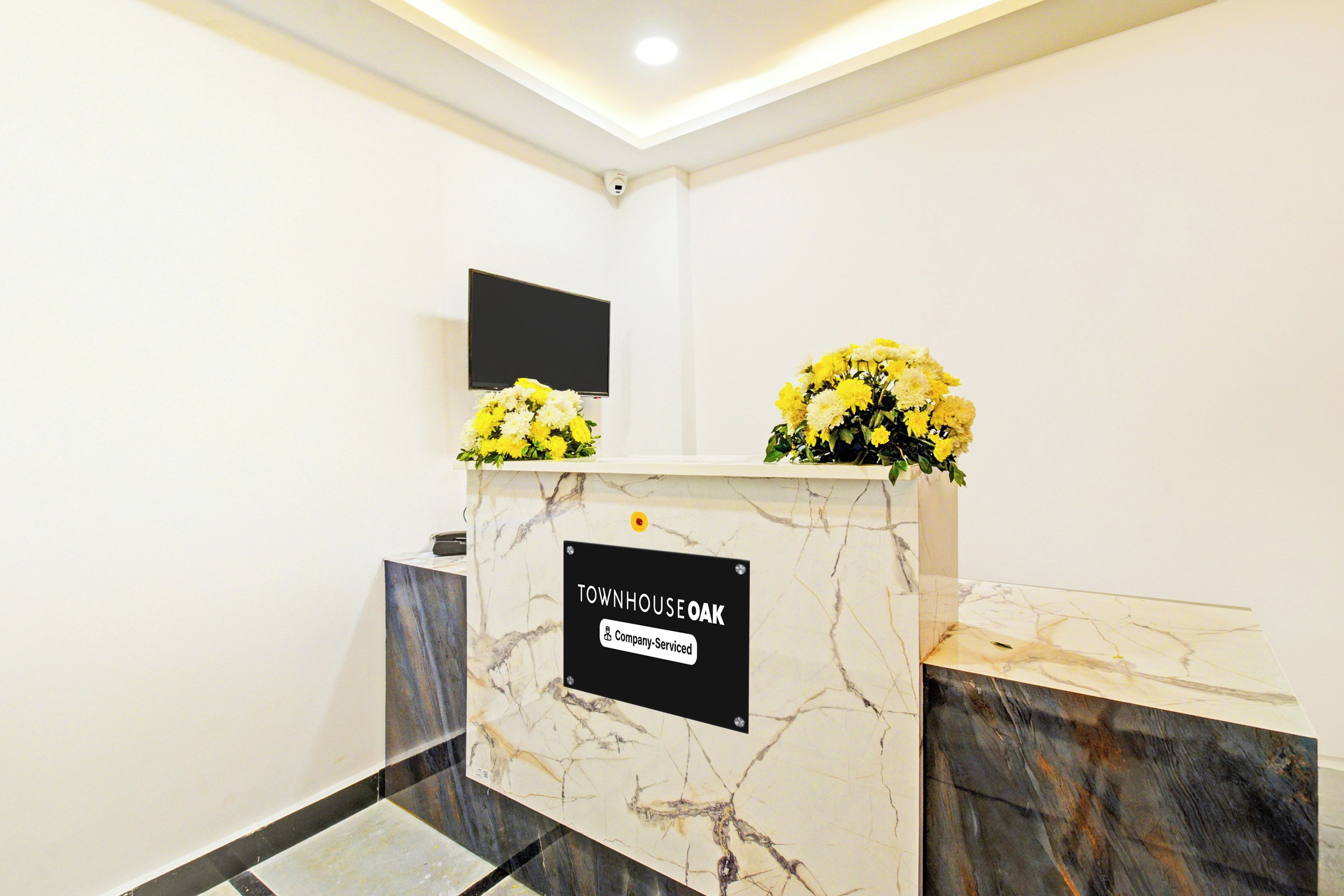 Un mostrador de recepción con un letrero que indica que es un área de servicio de empresa, un televisor de pantalla plana montado en la pared y un jarrón con flores amarillas sobre el mostrador.