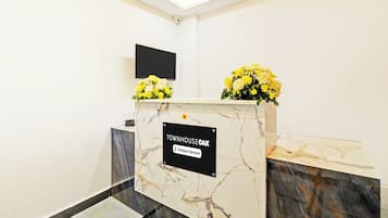 Un mostrador de recepción con un letrero que indica que es un área de servicio de empresa, un televisor de pantalla plana montado en la pared y un jarrón con flores amarillas sobre el mostrador.