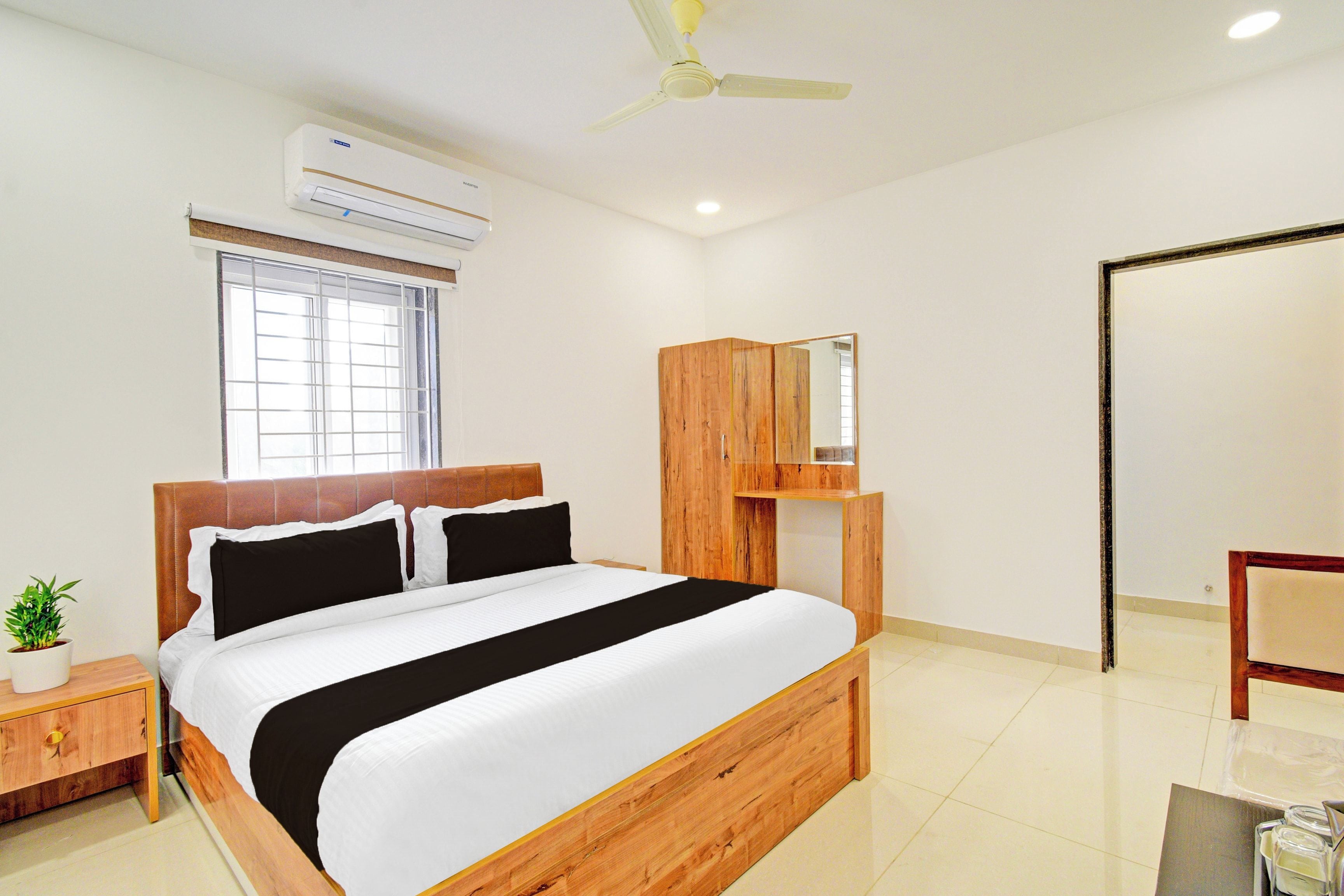 Habitación de hotel con cama, mesita de noche, planta en maceta, ventana, equipo de aire acondicionado, ventilador de techo y armario de madera.