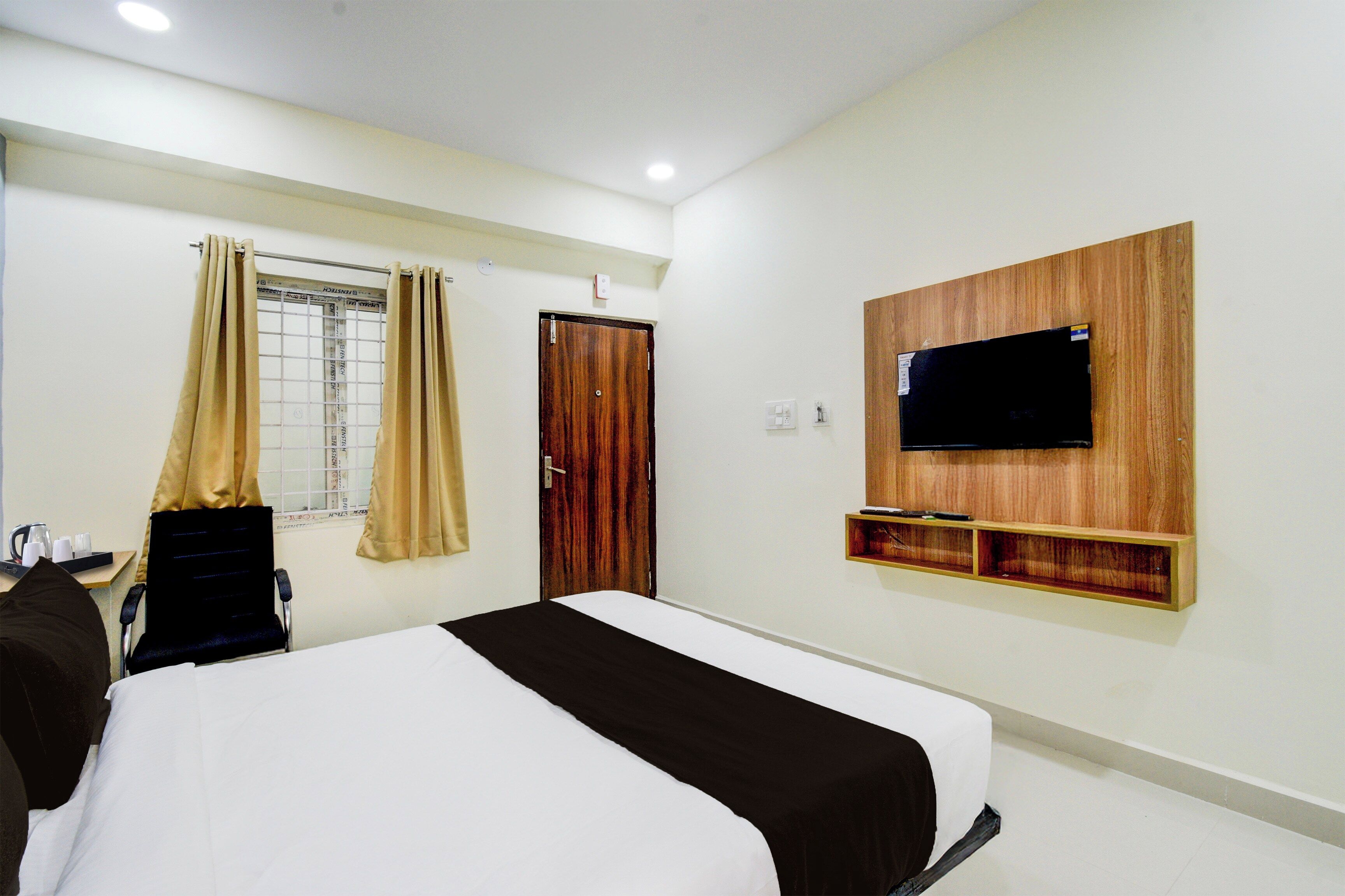 Habitación de hotel con cama, televisor montado en la pared, ventana con cortinas y estante de madera debajo del televisor.