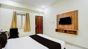 A hotel room with a bed, a TV mounted on the wall, a window with curtains, and a wooden shelf below the TV.