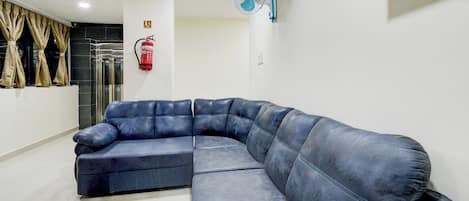 A modern living room with a large L-shaped sectional sofa, a ceiling fan, a fire extinguisher, and a wall-mounted TV.