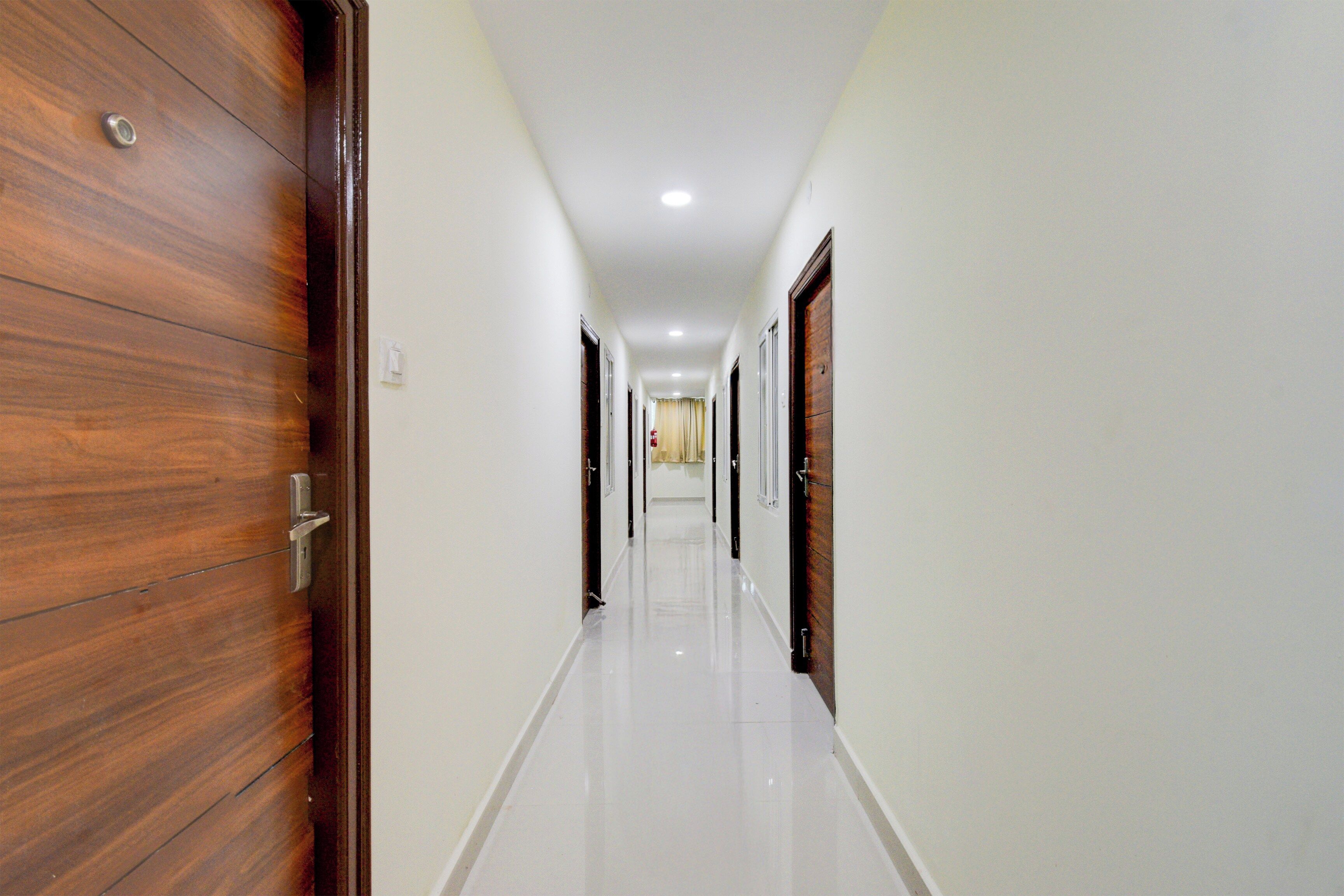 Un pasillo con puertas de madera a un lado y paredes blancas al otro, que conduce a una habitación con una ventana.