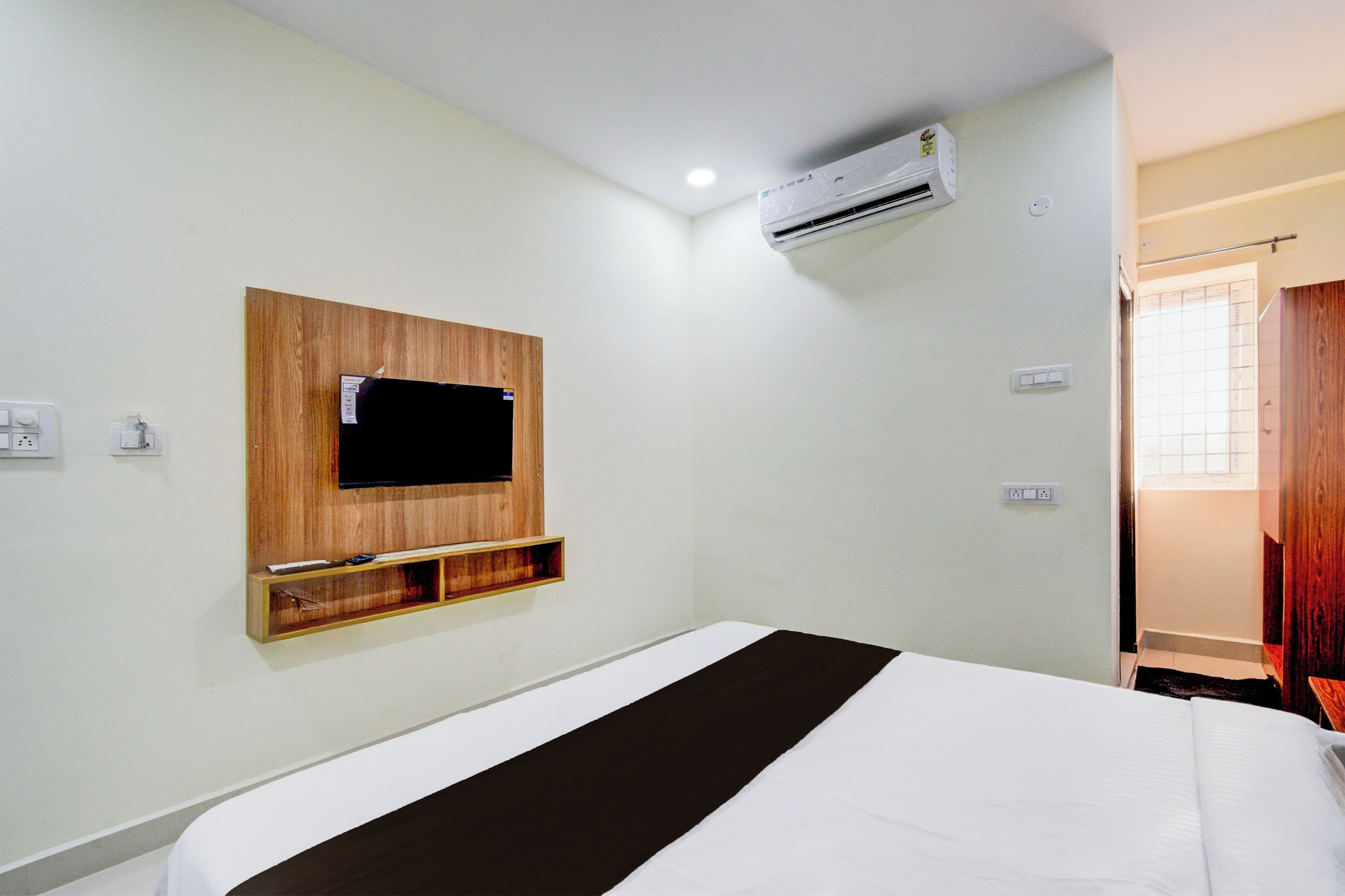 A hotel room with a bed, a mounted TV, an air conditioning unit, and a window with blinds.
