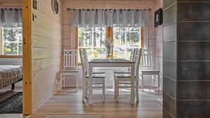 Interior de una cabaña de madera con una mesa de comedor, sillas, una ventana con cortinas y una chimenea.