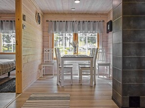 Interior de una cabaña de madera con una mesa de comedor, sillas, una ventana con cortinas y una chimenea.