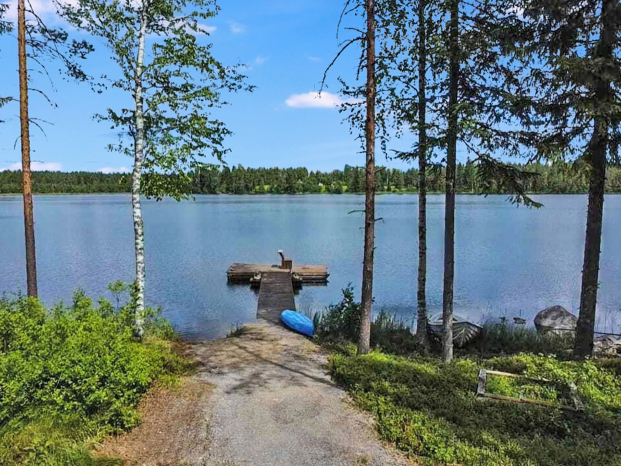 Järvi, jonka rannalla on laituri, joka ulottuu veden päälle. Järveä ympäröivät puut ja taivaalla on kirkas, selkeä sää.
