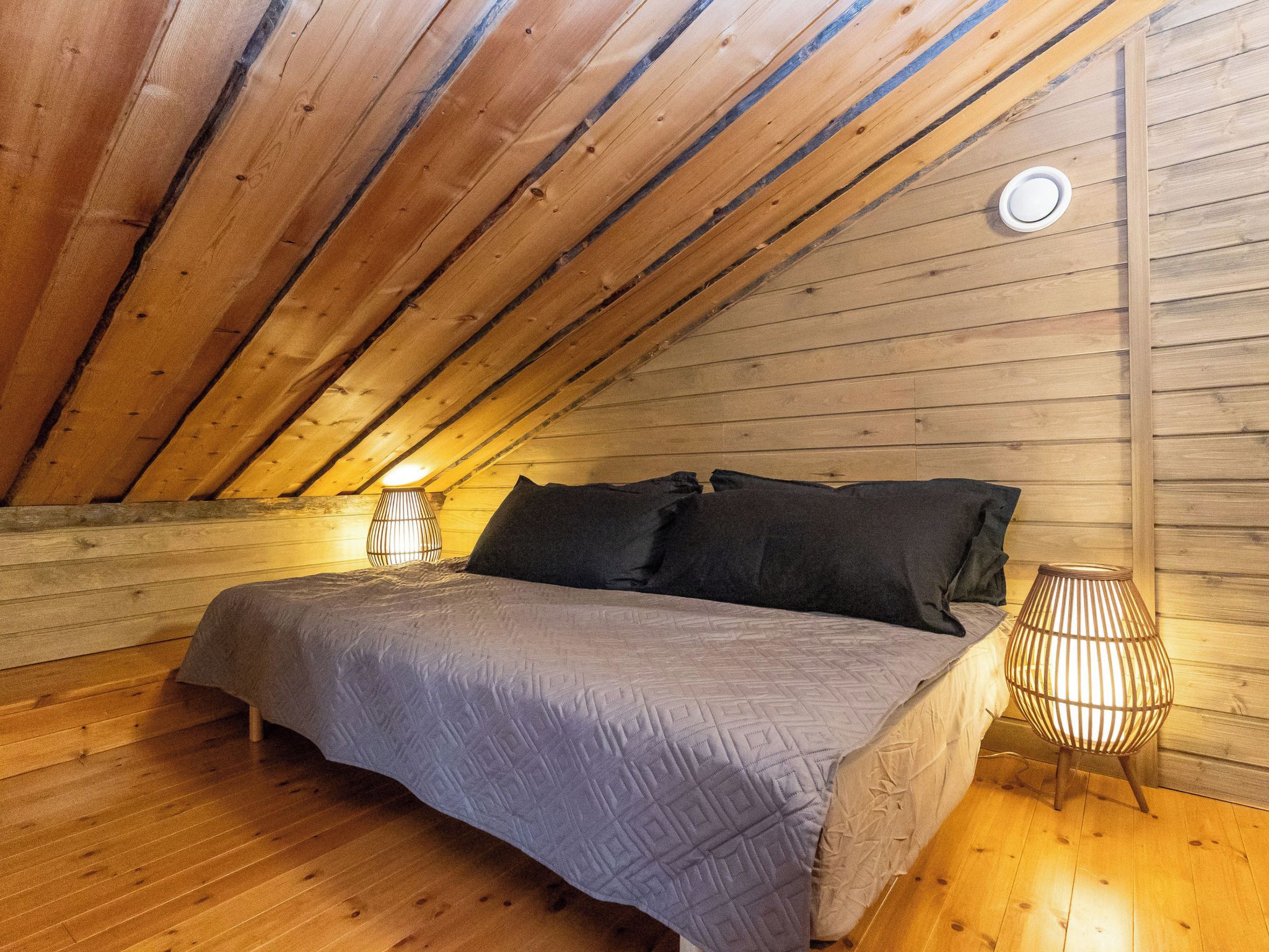 Ein Schlafzimmer mit schrägverdeckter Decke, Holzwänden, einem Bett mit grauer Tagesdecke, zwei schwarzen Kissen und einer Stehlampe.