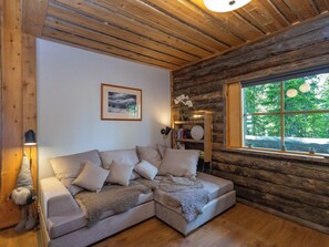 A cozy cabin interior with a sectional sofa, a bookshelf, a framed picture, and a window with a view of greenery. - Honka 1a by Interhome (Kuusamo)