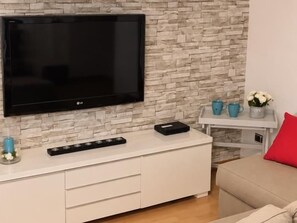 Un salon moderne avec un téléviseur à écran plat fixé sur un mur imitant la pierre, un meuble de rangement blanc avec des tiroirs, une petite table d’appoint agrémentée d’objets décoratifs et un coussin rouge sur un canapé beige.