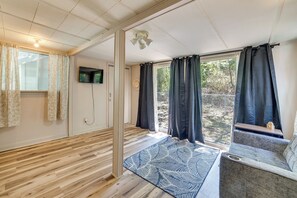 A room with a wooden floor, a flat-screen TV mounted on the wall, a window with curtains, and a view of the outdoors. - Cedar Creek Lake House! Beautiful Lake Views! (Mabank)