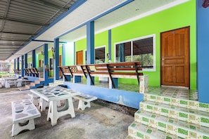 A covered outdoor seating area with benches and tables, a covered walkway with a tiled floor, and a building with green and blue walls and wooden doors. - Kwanjai resort (Tha Mai)