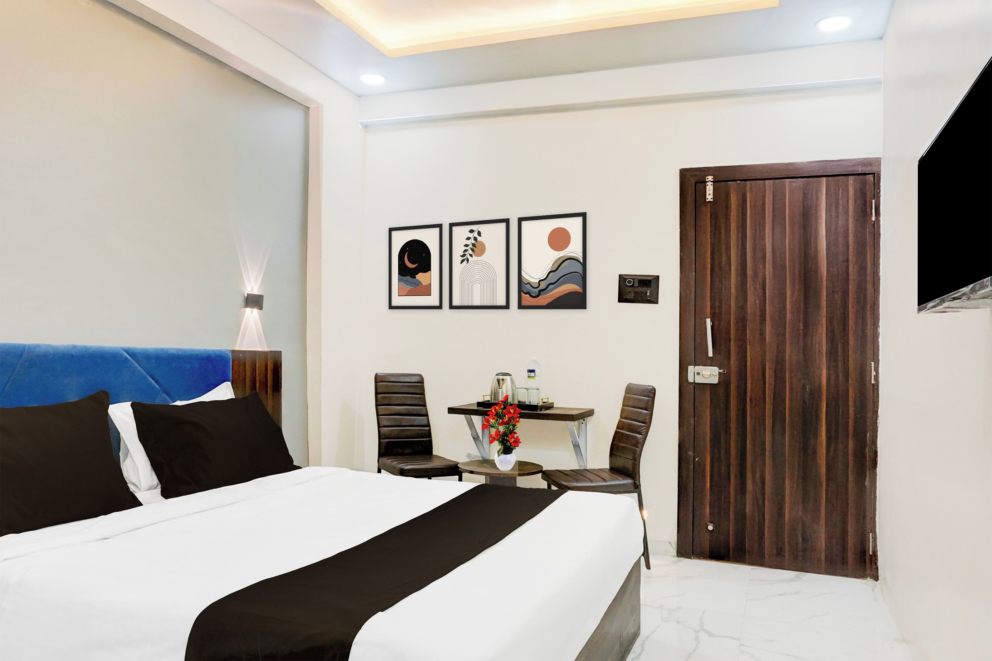 Habitación de hotel con cama, escritorio y sillas, mesa con un jarrón de flores y una puerta.