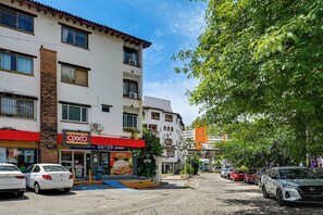 A street view with a two-story building featuring a red awning, a visible store with a sign, parked cars, and a tree-lined sidewalk. - Walk Everywhere! Central Puerto Vallarta Condo (Puerto Vallarta)