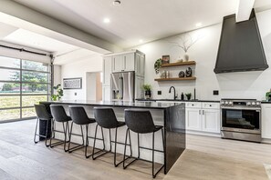 A modern kitchen with a large island, bar stools, stainless steel appliances, and a view through the glass door. - Golf Sim & Sports Arcade! Luxe Indy Escape (Indianapolis)