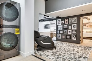 A laundry room with a washing machine and dryer, a seating area with a chair, and a wall with framed pictures. - Golf Sim & Sports Arcade! Luxe Indy Escape (Indianapolis)
