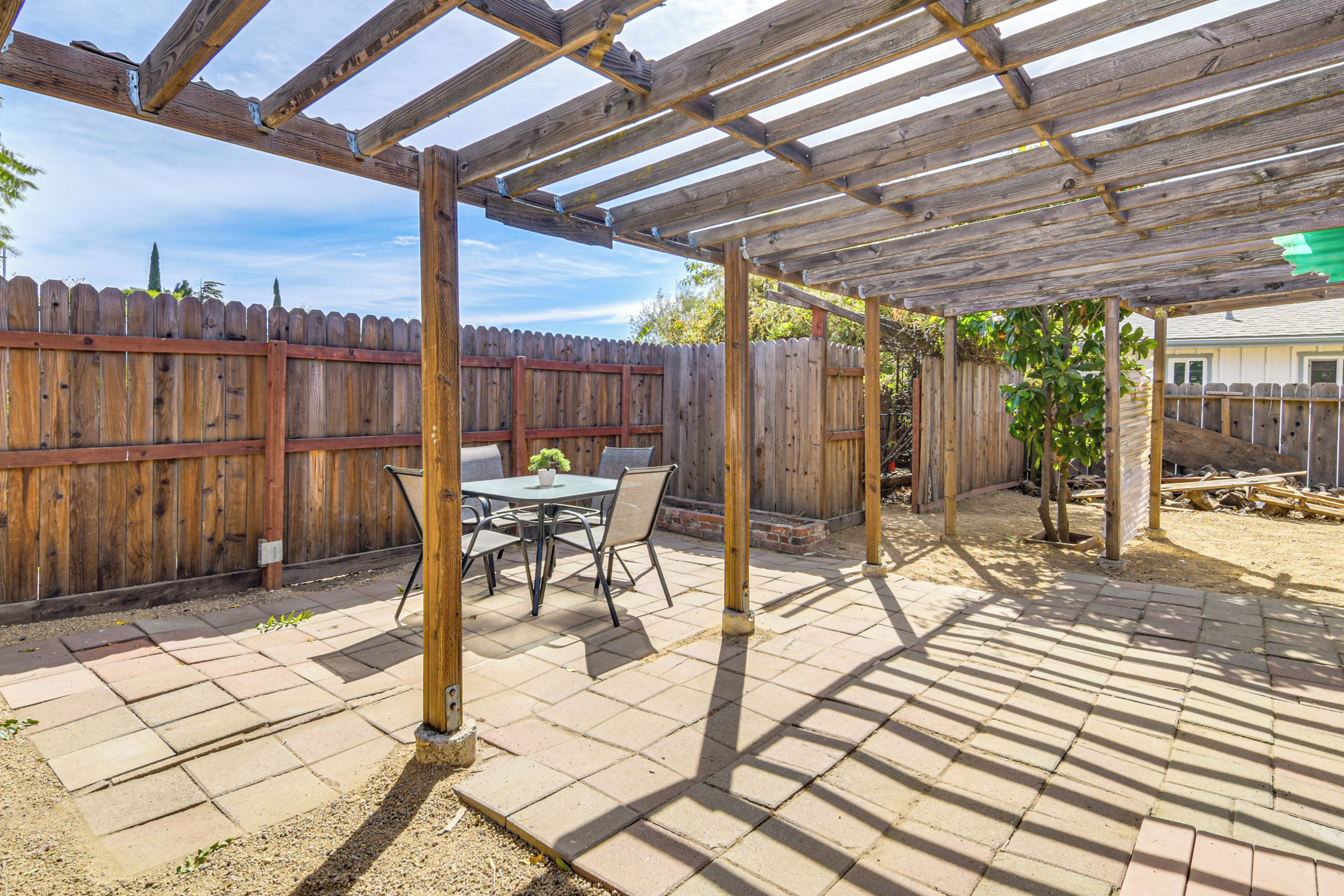 A covered patio area with a wooden pergola, a table with chairs, and a wooden fence in the background.