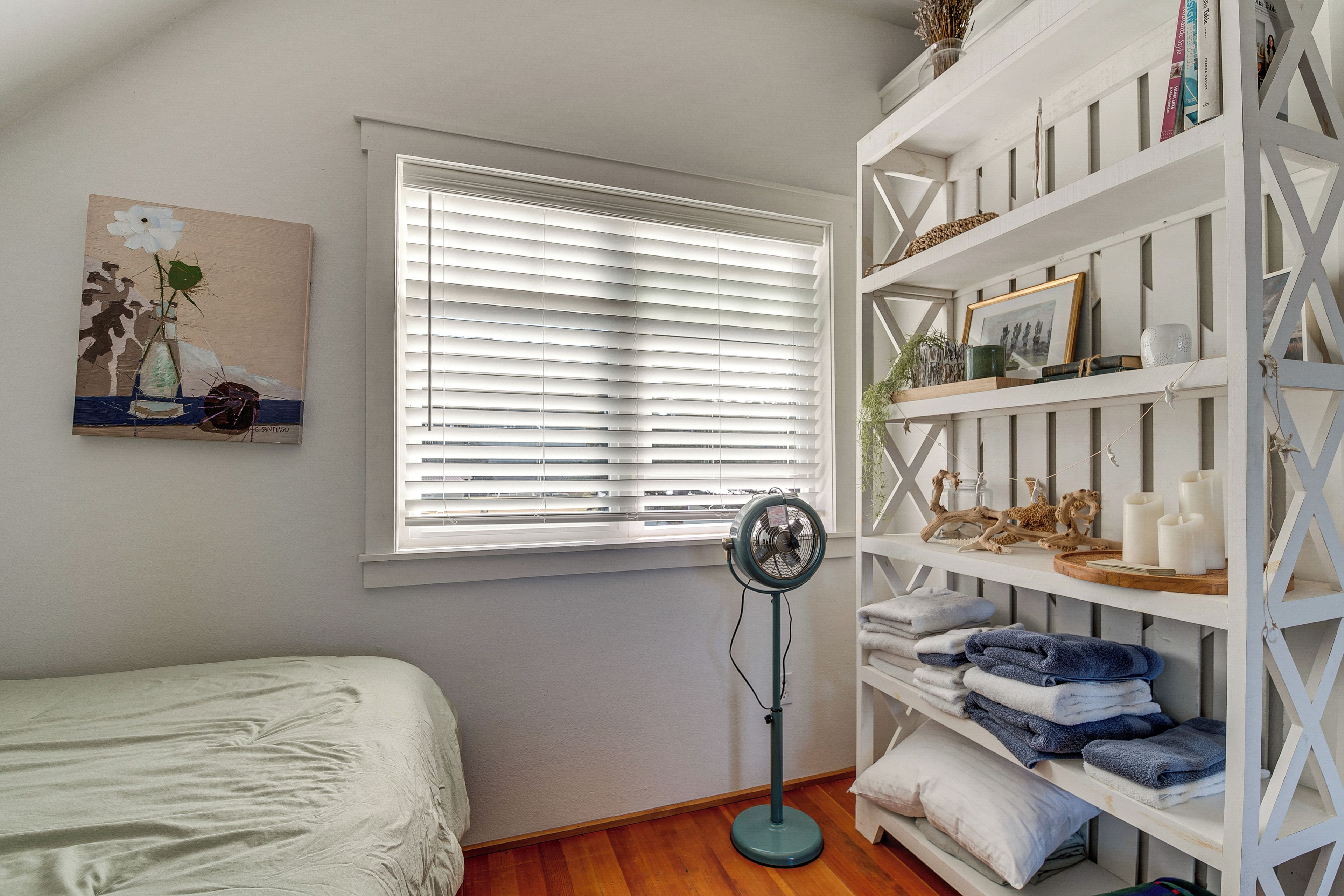 A bedroom with a bed, a standing fan, a window with blinds, and a bookshelf with various items.