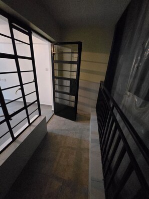 Corredor com corrimão de metal preto, porta com painel de vidro e piso ladrilhado.