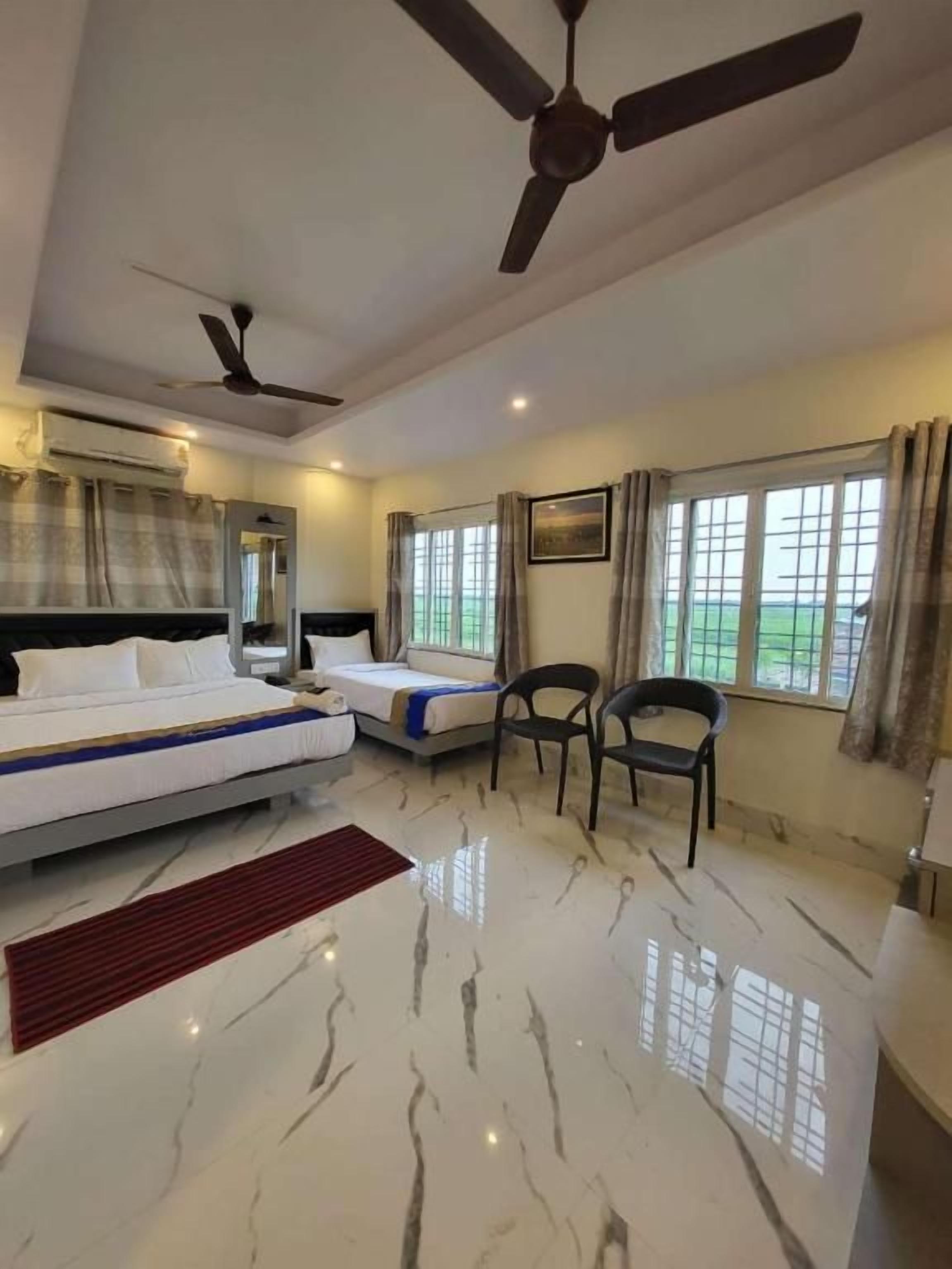 Habitación de hotel con cama, dos mesitas de noche, una silla, un escritorio, ventana con cortinas y ventilador de techo.