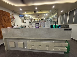 A hotel reception desk with a digital clock displaying the time, a reception sign, and various informational posters. - HOTEL SHREESH MAYAPUR (Krishnanagar)