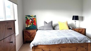Un dormitorio con una cama de madera, una mesita de noche con una lámpara, un aparador y un cuadro colorido en la pared.